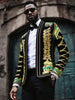 Man wearing a ornate jacket with gold and green embroidery against a stone wall.