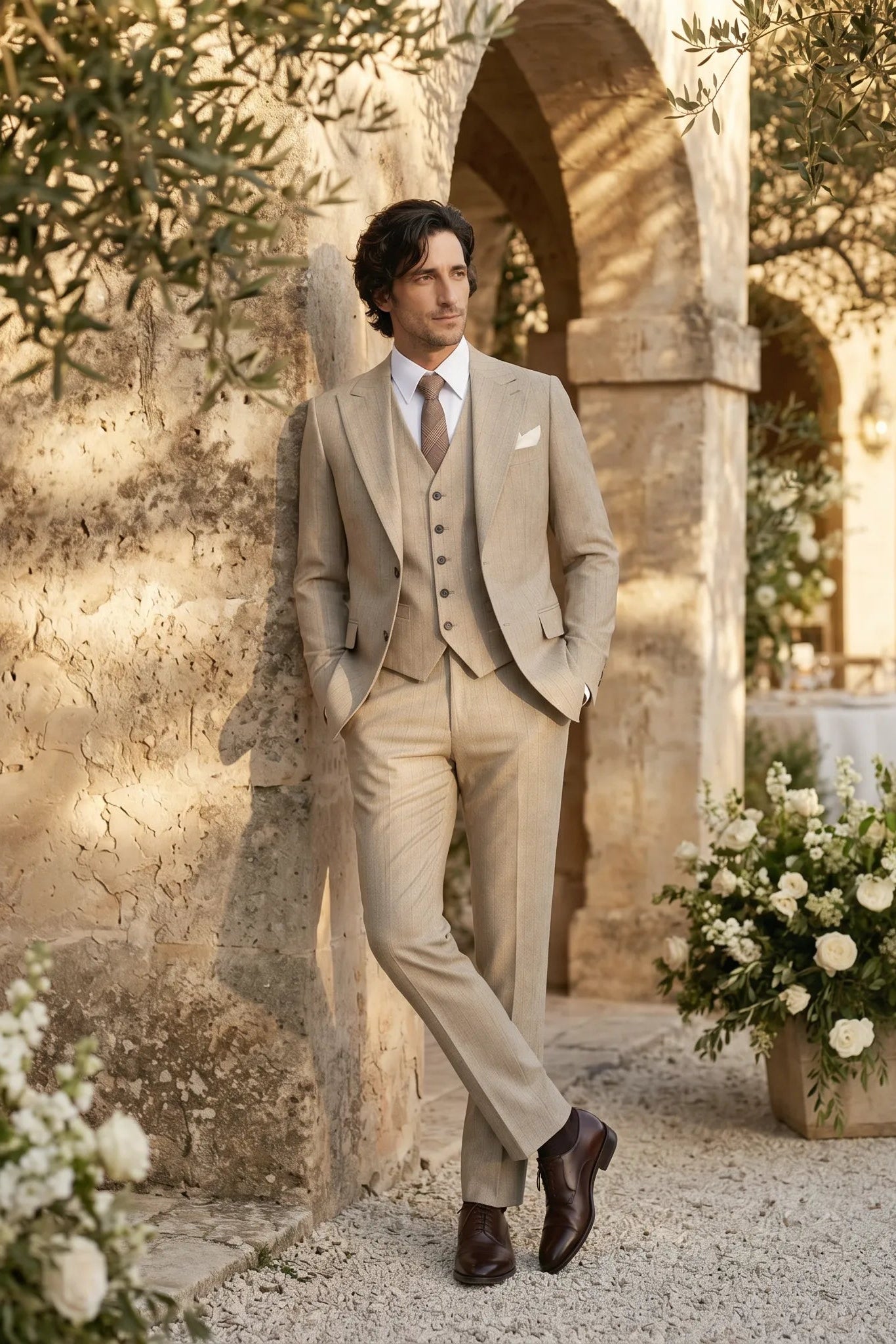 Man in a beige suit standing in an outdoor setting with floral arrangements and stone architecture.
