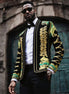 Man wearing a ornate jacket with gold and green embroidery against a stone wall.