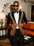 Man in a black and gold embroidered suit standing in an elegant room with orange sofa and decorative items.
