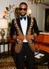 Man in a black and gold embroidered suit standing in an elegant room with orange sofa and decorative items.