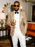 Man in a gold patterned jacket and white pants holding a glass in an elegant room.