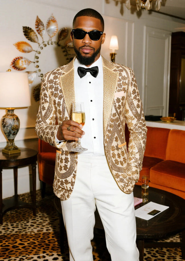 Man in a gold patterned jacket and white pants holding a glass in an elegant room.