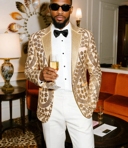 Man in a gold patterned jacket and white pants holding a glass in an elegant room.